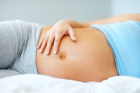 A pregnant woman gently rests her hand on her rounded belly while lying on a soft bed, conveying a sense of comfort and anticipation.