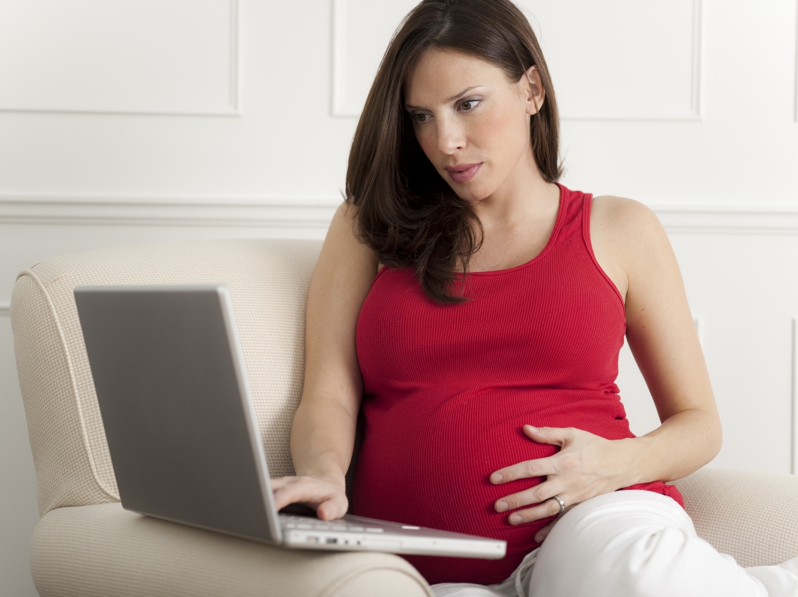 A pregnant woman seated on a couch, wearing a red tank top, gently cradles her belly while working on a laptop.