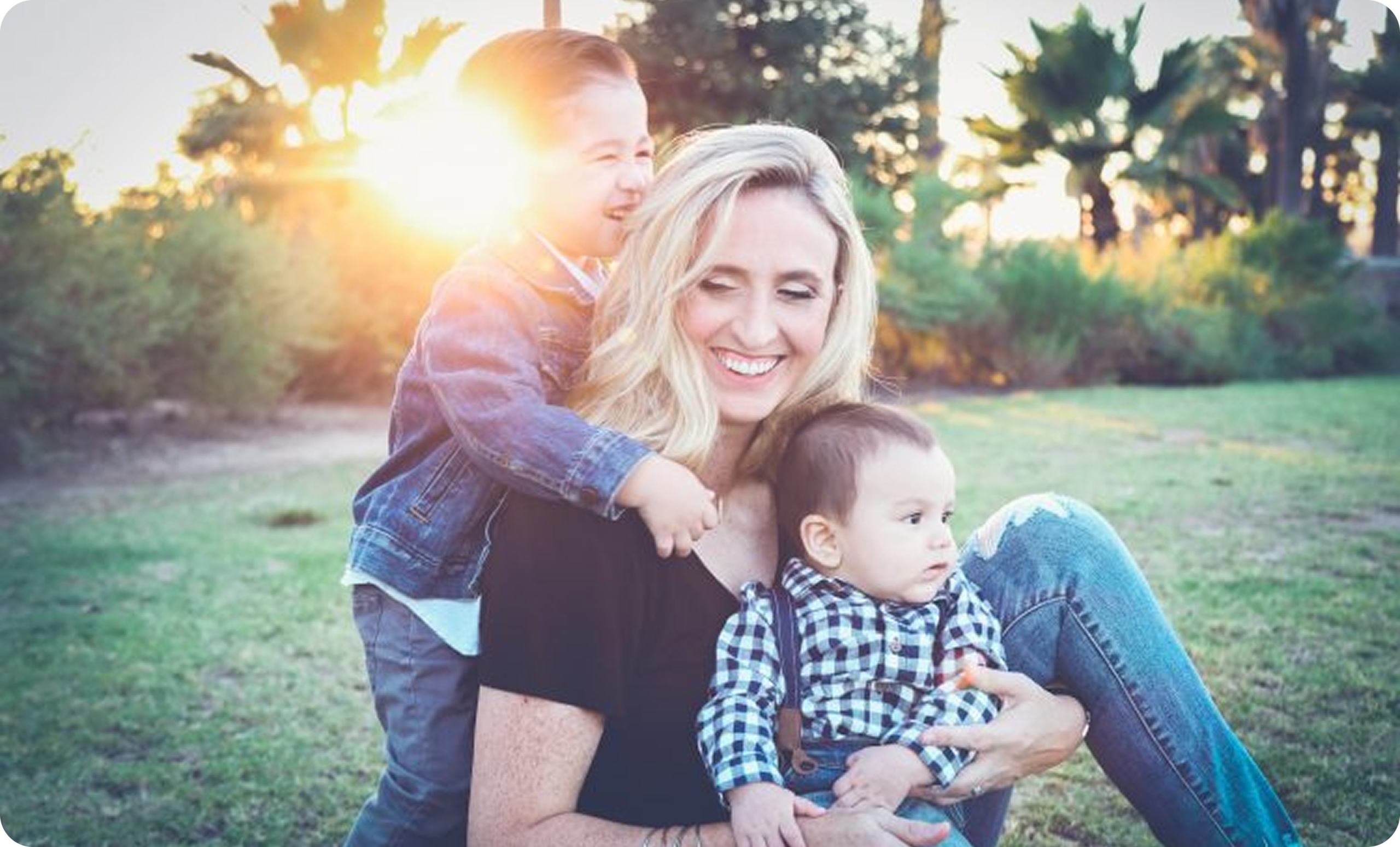 A woman sits on grass with two children, enjoying sunset lights and surrounded by lush greenery.