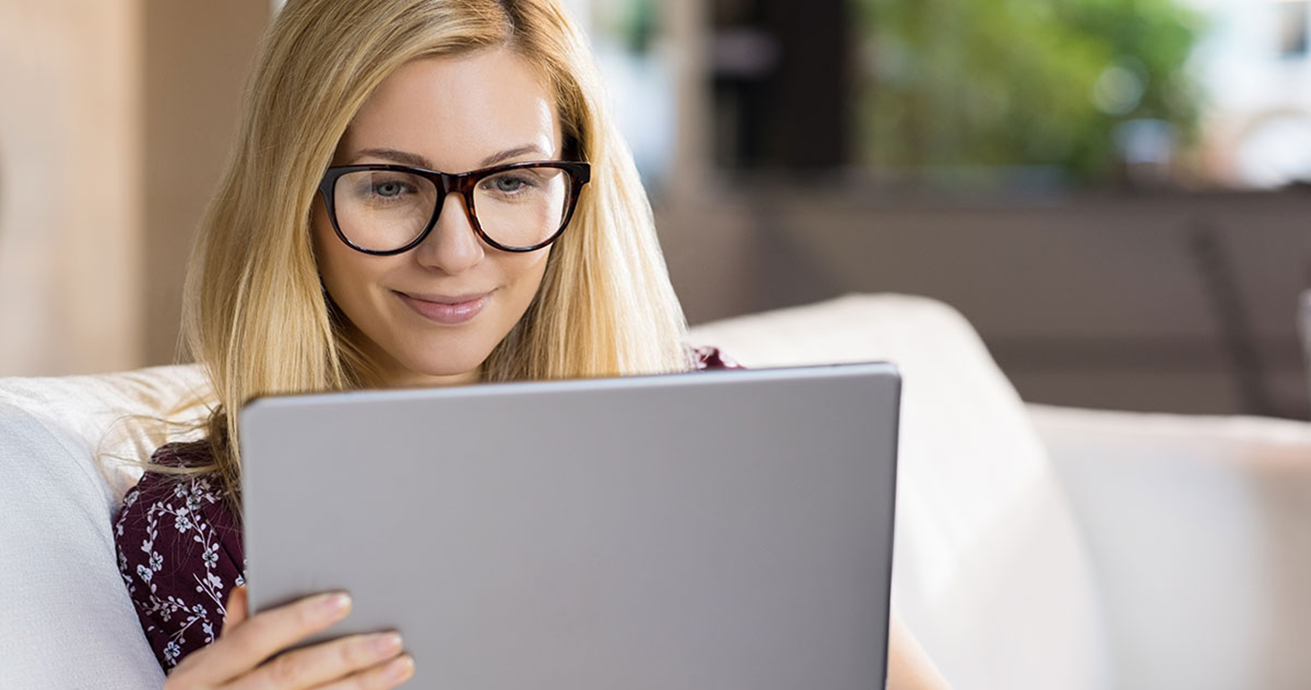 A person with long blonde hair sits on a couch, intently using a tablet in a cozy, softly lit environment.