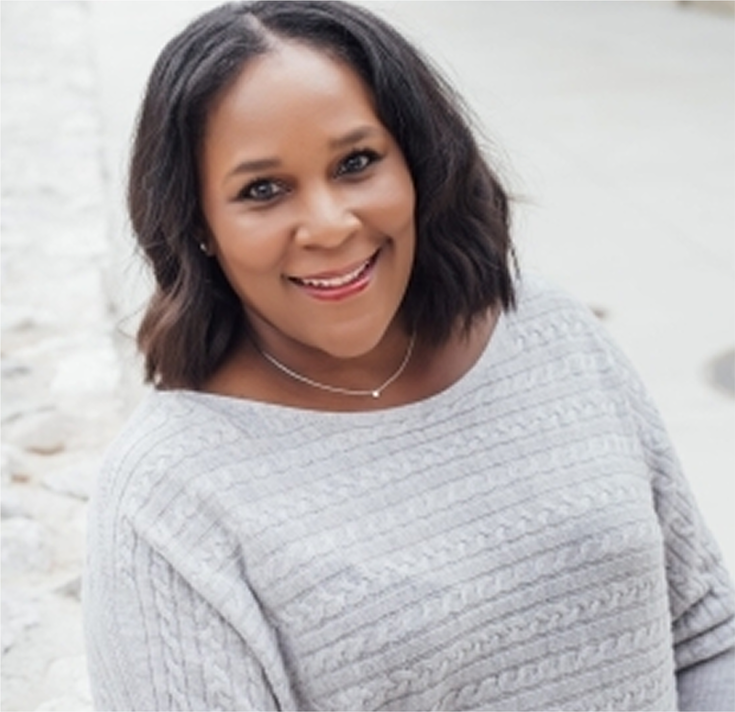 A woman poses casually outdoors, wearing a cozy gray sweater and a delicate necklace, with a stone wall in the background.