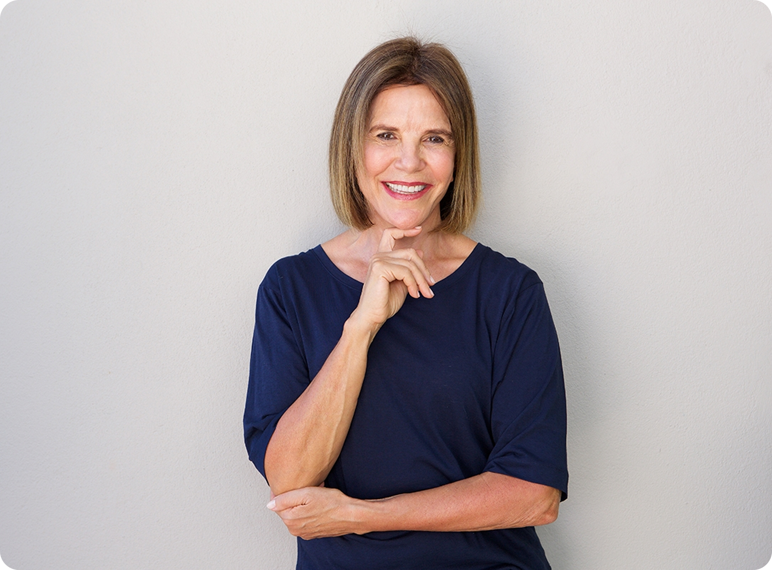 A woman with shoulder-length hair, wearing a navy blue shirt, poses with her finger to her lips, suggesting silence or contemplation against a beige wall.