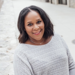 A woman poses casually outdoors, wearing a cozy gray sweater and a delicate necklace, with a stone wall in the background.