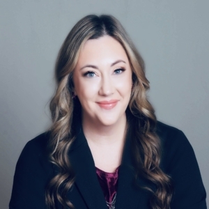 A person with wavy hair wearing a dark blazer and a burgundy top, set against a simple gray background.