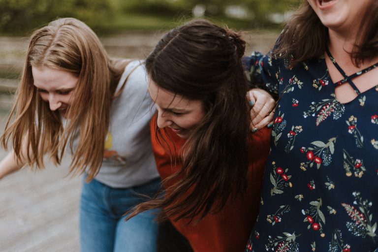 Three women enjoying a playful moment together outdoors, laughing and wearing casual attire, surrounded by greenery.
