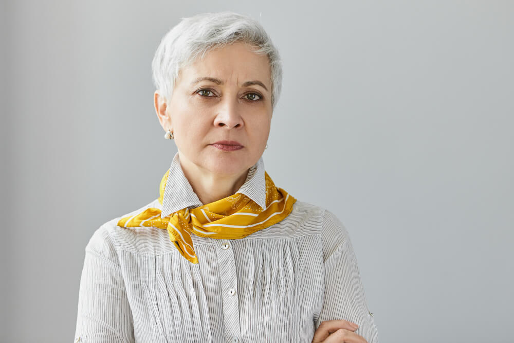 A woman with short gray hair, wearing a striped shirt and a yellow scarf, stands with her arms crossed in front of a light gray background.