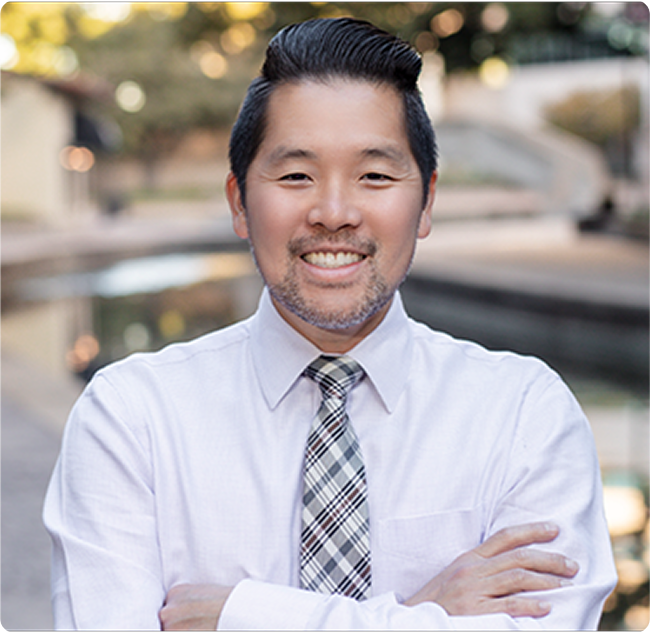A man in a white dress shirt and plaid tie stands confidently with arms crossed, against a blurred outdoor background.