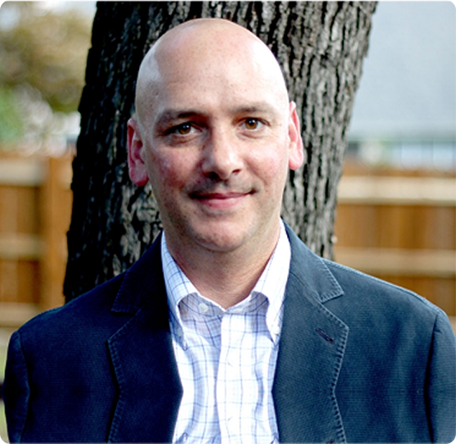 A person wearing a light blue shirt and dark blazer stands in front of a tree, with a wooden fence in the background.