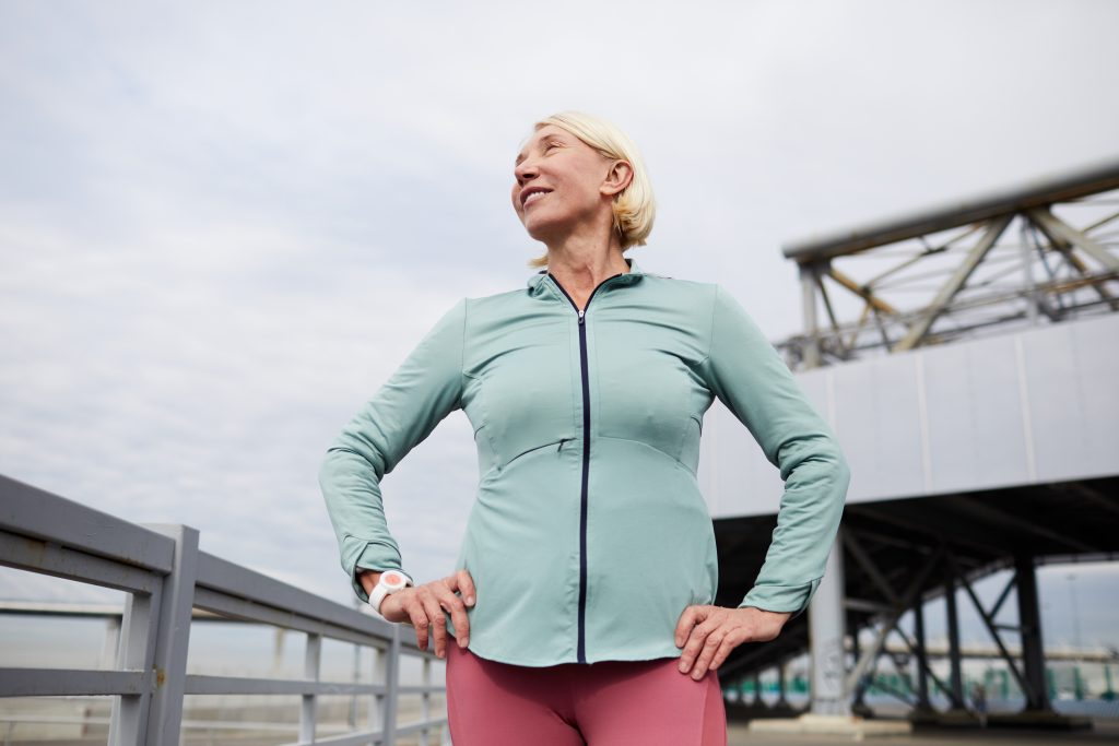 Mature happy sportswoman in activewear keeping her hands on hips while exercising or having short break after workout