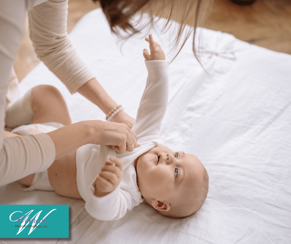 A baby lying on a blanket as their mother dresses them.